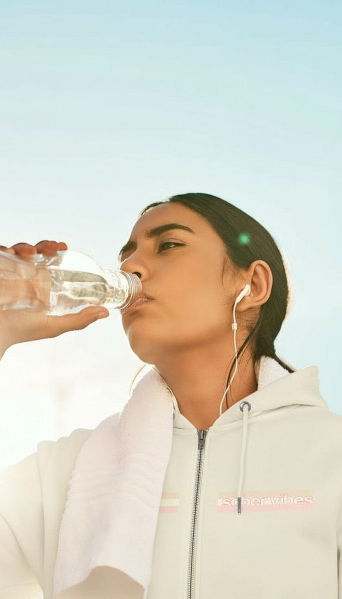 Women with earbuds drinking water wearing a stripped cream sober vibes zip-up hoodie